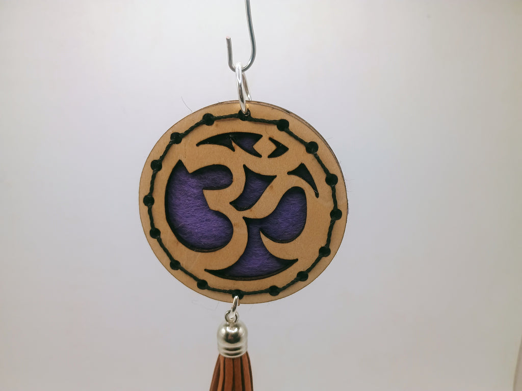 Decorative hanging ornament with Om symbol over purple felt and reddish tassel on a plain background