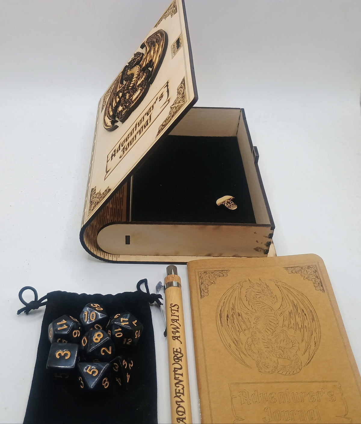 A themed tabletop gaming set featuring a miniature book-shaped box with an engraved cover, a black velvet pouch containing polyhedral dice, a tan notebook, and a wooden pen